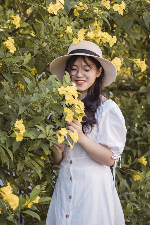 免費 白色的裙子和白色的帽子，站在黃色的花樹旁邊的女人 圖庫相片