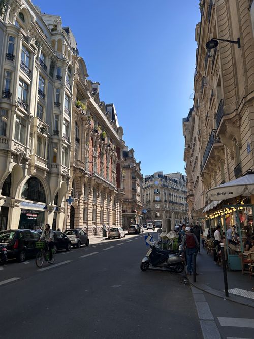 Free A bustling Paris street with cafes, historic buildings, and a cyclist under a clear blue sky. Stock Photo