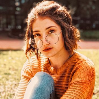 Portrait of a young woman with eyeglasses sitting in a sunlit park, wearing a cozy sweater.