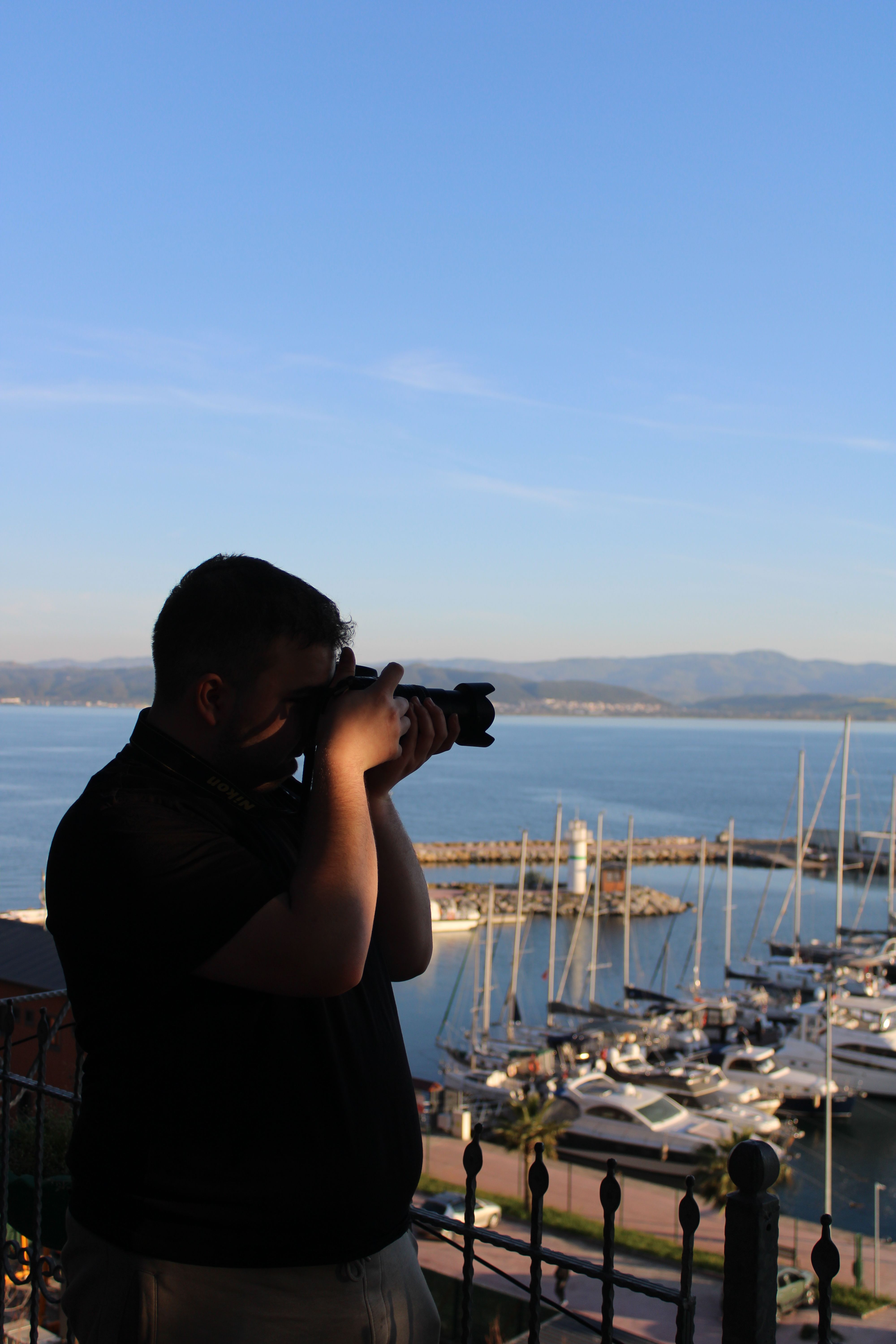 A photographer takes a picture of a marina at sunrise with yachts and mountains in the background.