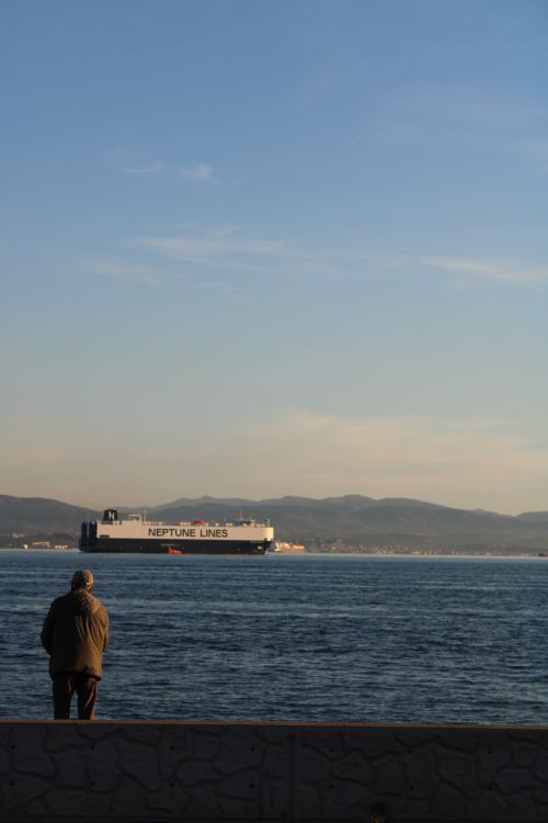 Free A senior stands by the sea observing the Neptune Lines ship with distant mountains in the background. Stock Photo