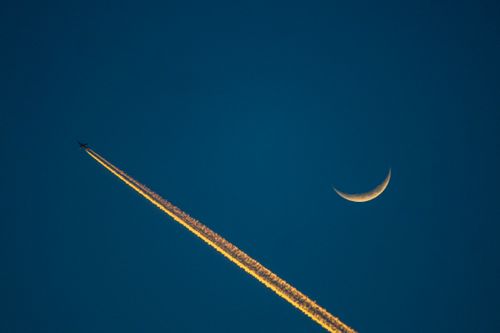 Free Airplane leaves contrail beside a crescent moon against deep blue night sky. Stock Photo
