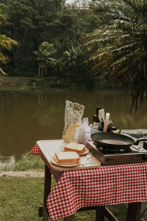 Free Outdoor breakfast preparation by a serene riverside campsite with a portable stove and sandwiches on a checked tablecloth. Stock Photo