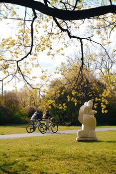 Two cyclists ride past a sculpture in a vibrant spring park scene.