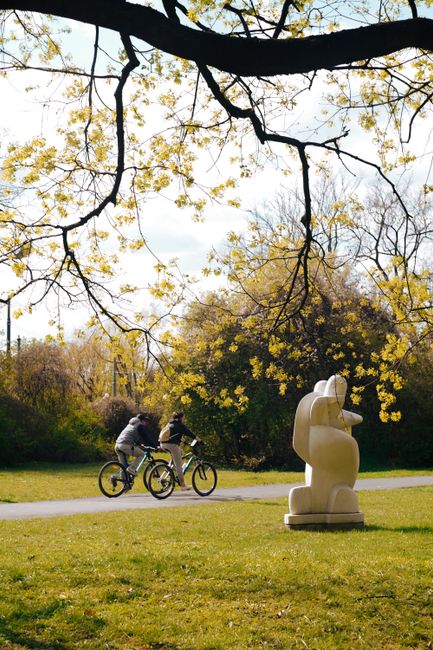 Two cyclists ride past a sculpture in a vibrant spring park scene.