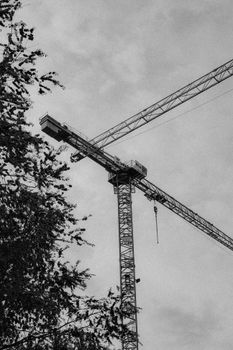 A monochrome view of a construction crane against a cloudy sky, with tree branches.