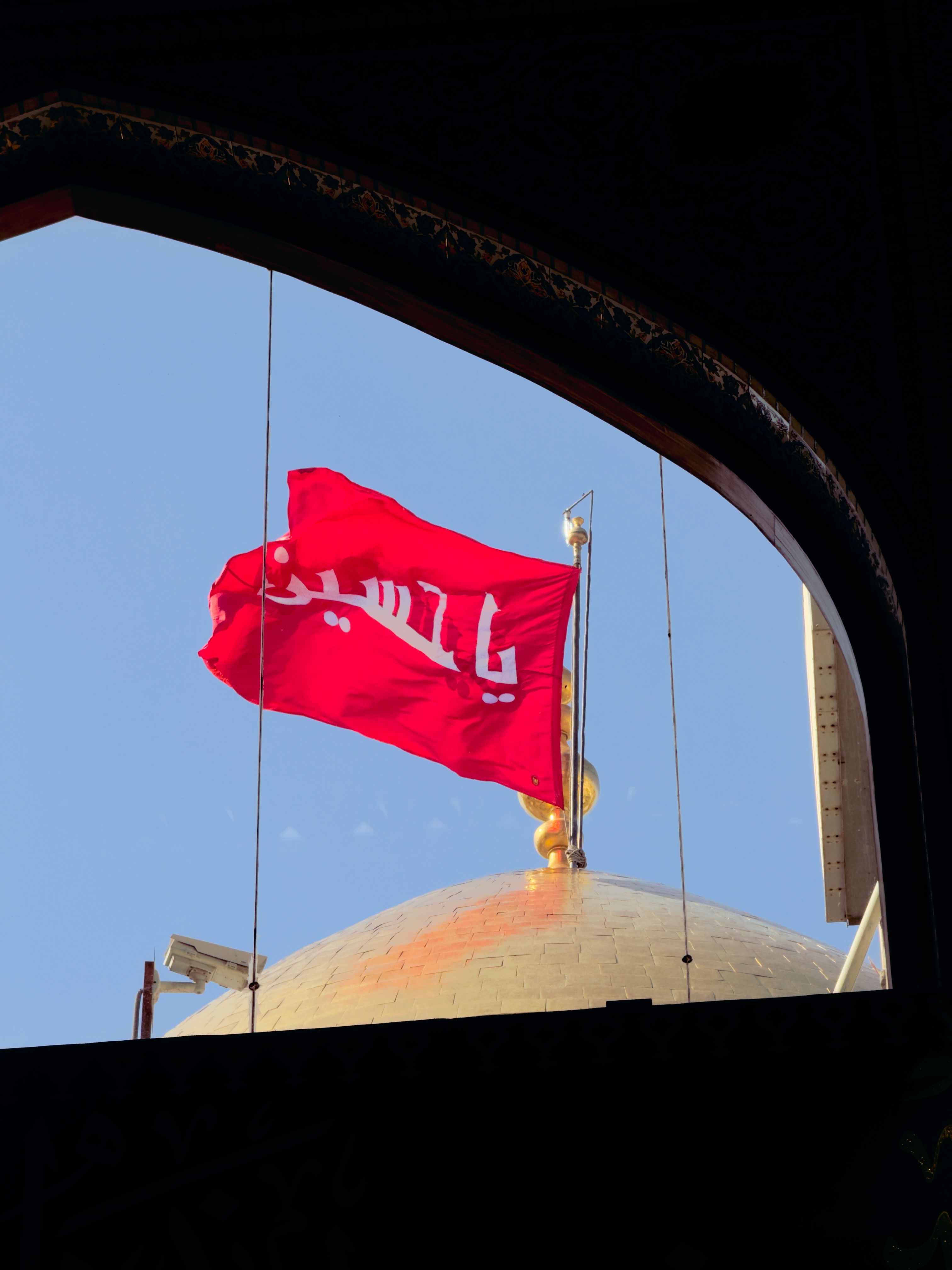 Red flag with Arabic script flies over golden dome of an Islamic shrine, viewed through arch.