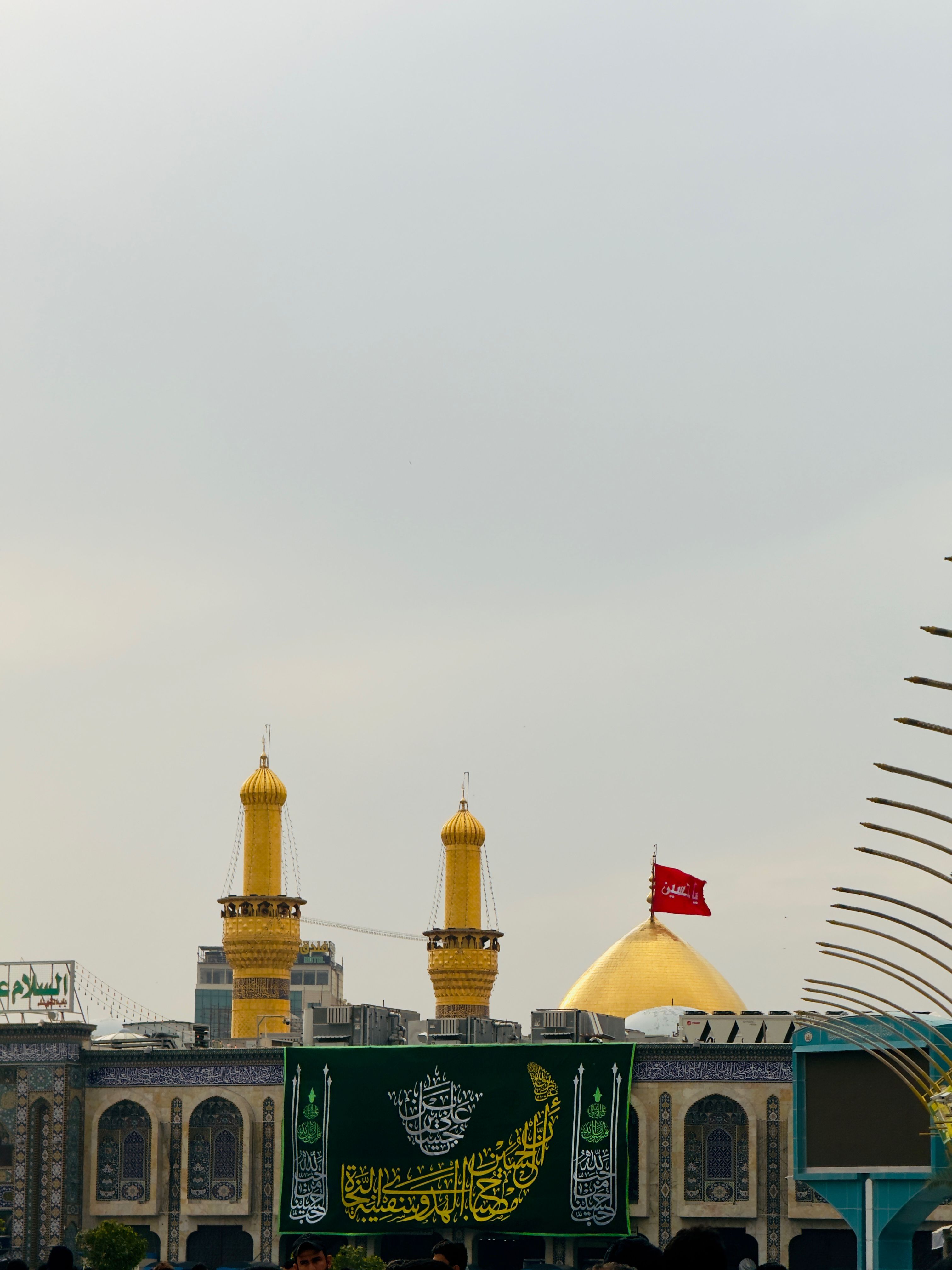 A view of the gold-domed shrine in Karbala, Iraq, with a red flag and Arabic inscriptions.