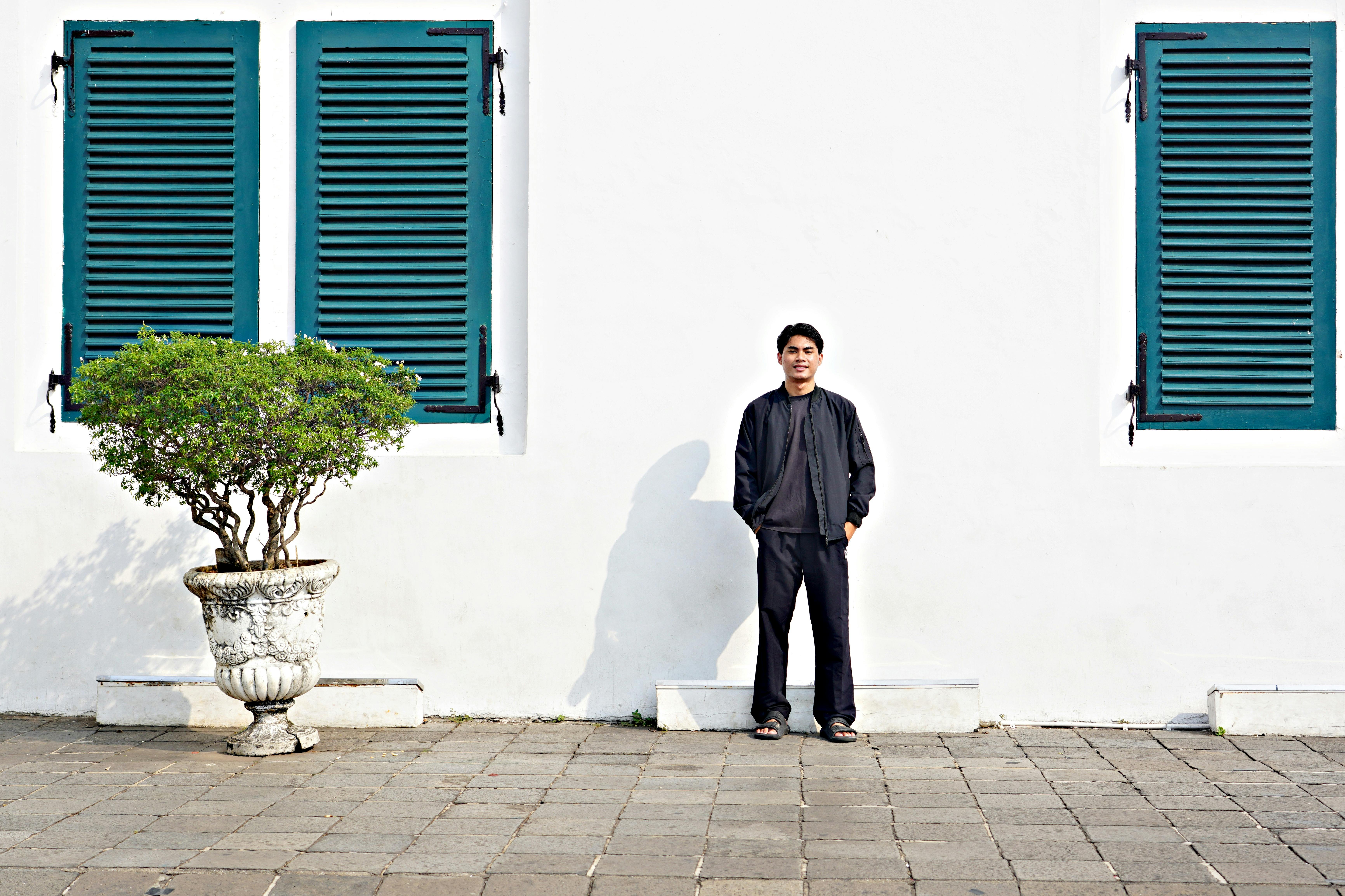 A man stands against a white wall with two green shuttered windows and a potted plant.