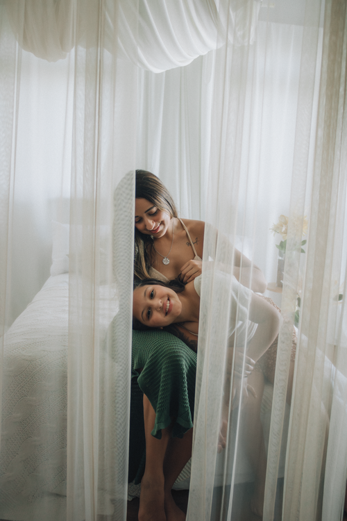 Free A loving mother and daughter share a tender moment behind sheer curtains in a serene bedroom setting. Stock Photo