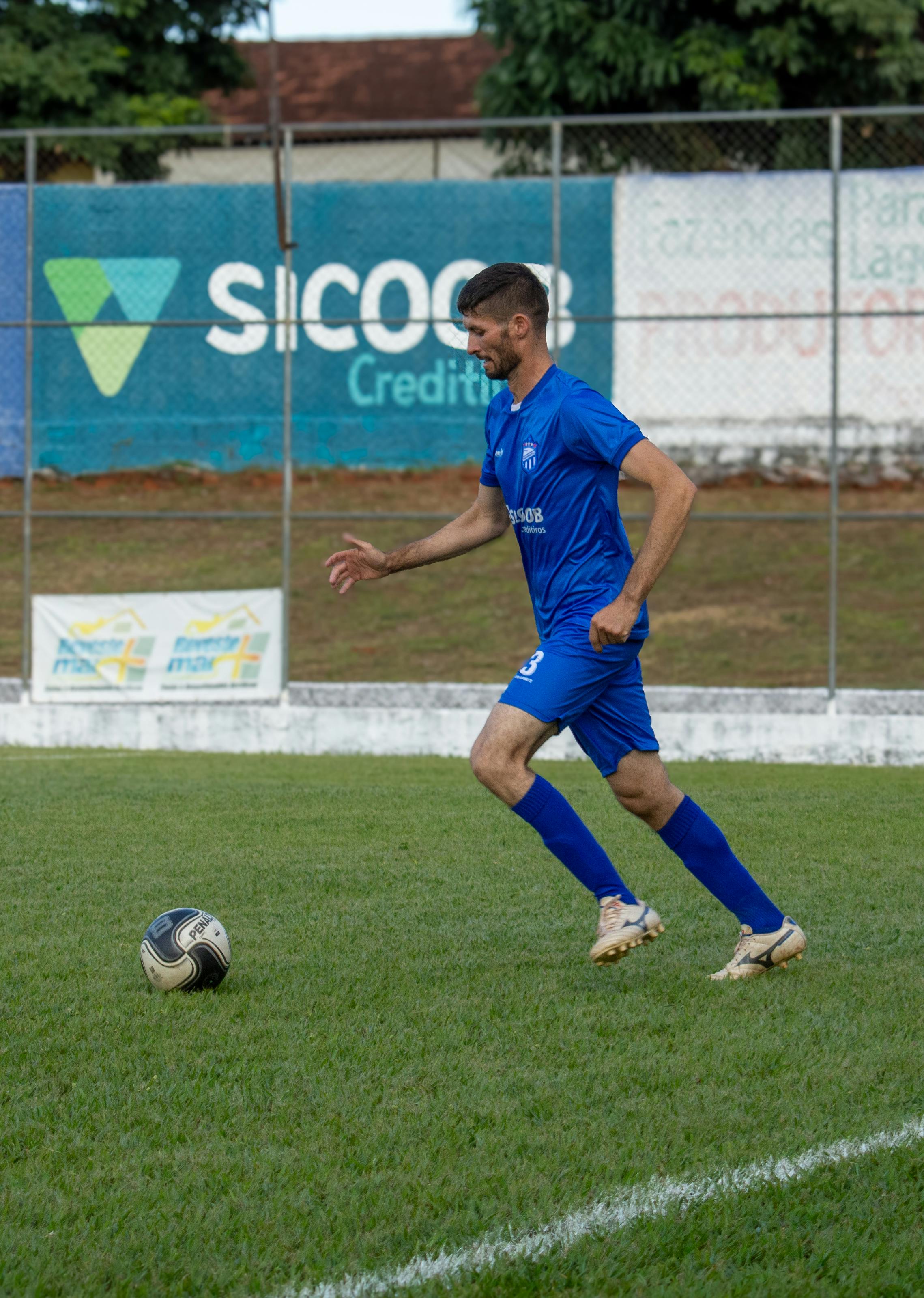 Focused soccer player in blue uniform dribbling during a daytime match on a grassy field outdoors.