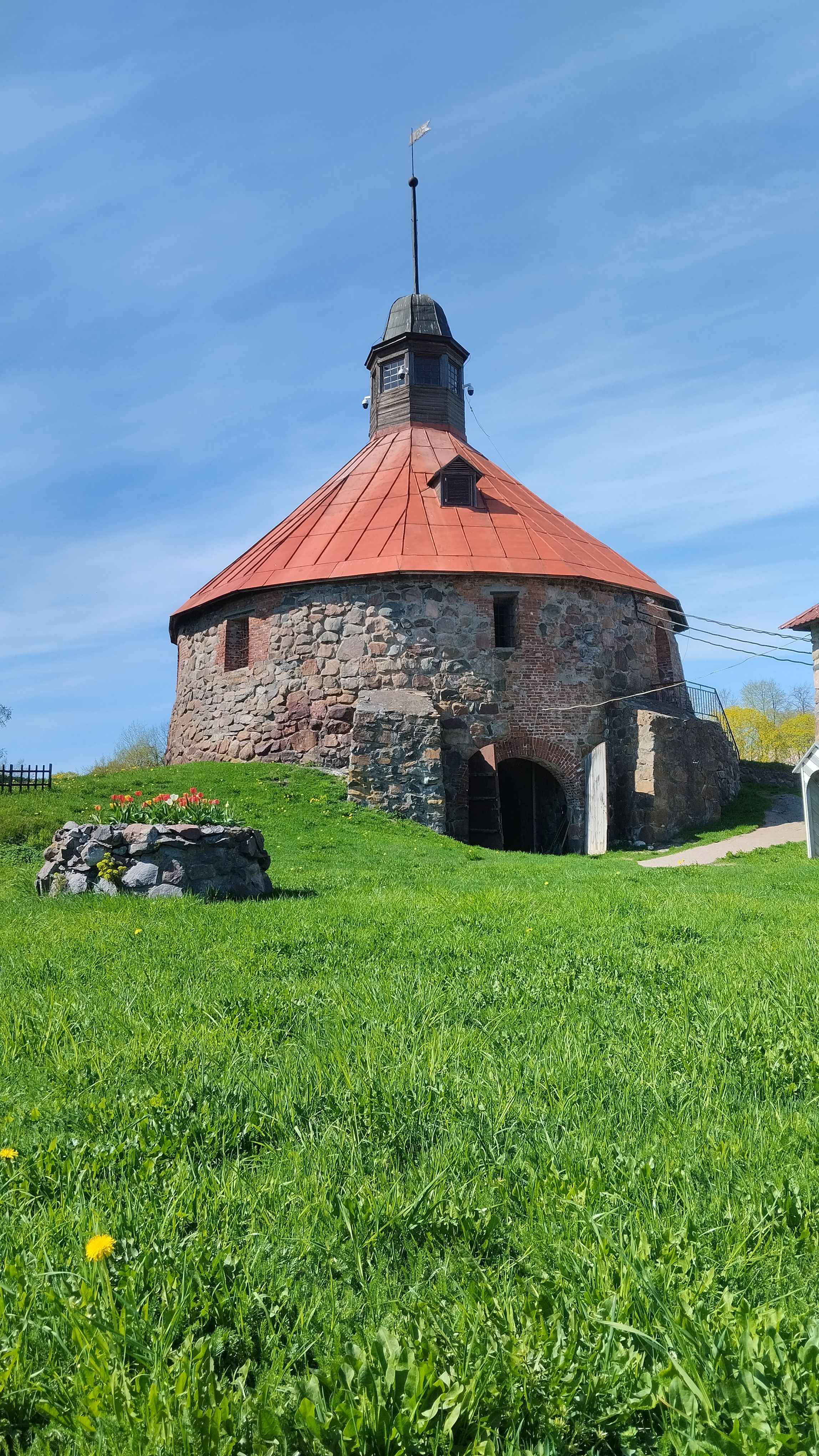 A vintage stone tower with a red roof amidst a green field under a blue sky.