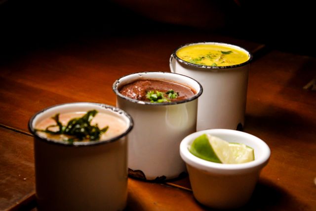 Three enamel cups of assorted soups with herb garnish beside a dish of lime wedges