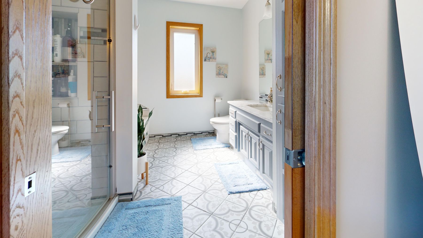 View into a bright bathroom with a long grey vanity, glass shower, patterned floor and a small timber-framed window