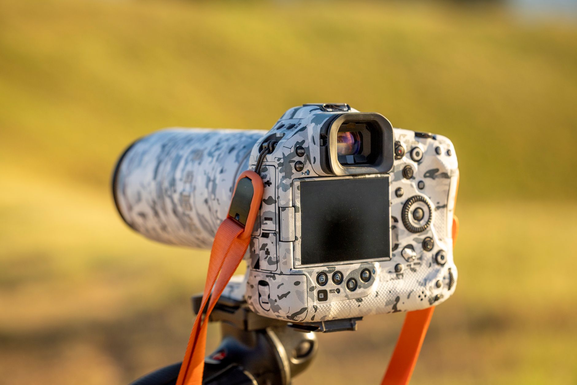 Camouflage-patterned DSLR camera with a telephoto lens on a tripod, in a sunny outdoor setting.
