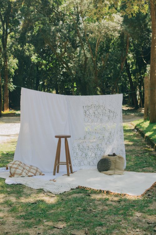 Free Cozy outdoor setup with white fabric, stool, and picnic blanket in a sunny forest setting. Stock Photo