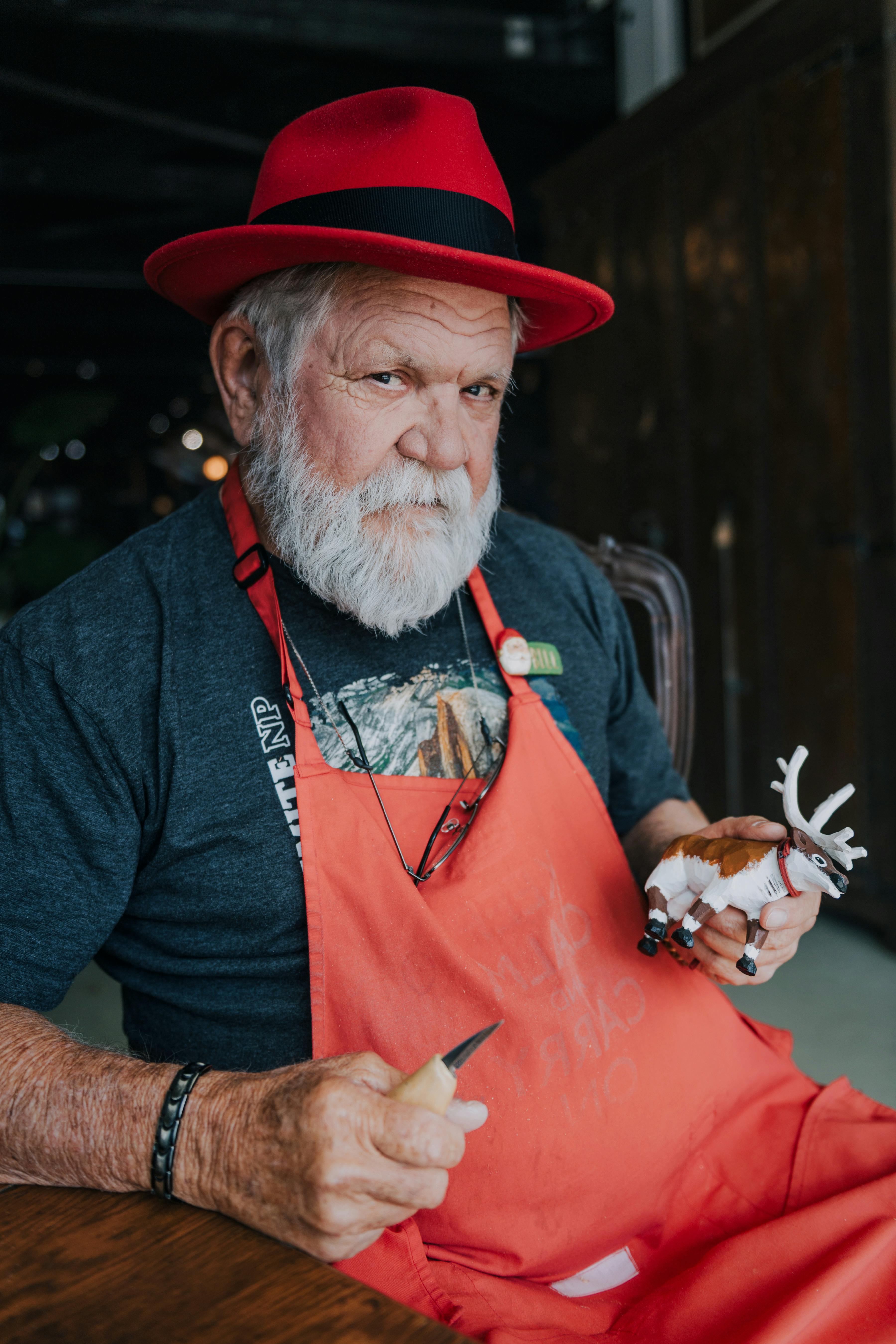 Senior craftsman in a red hat and apron carves a reindeer figurine indoors. Los Alamos, CA.