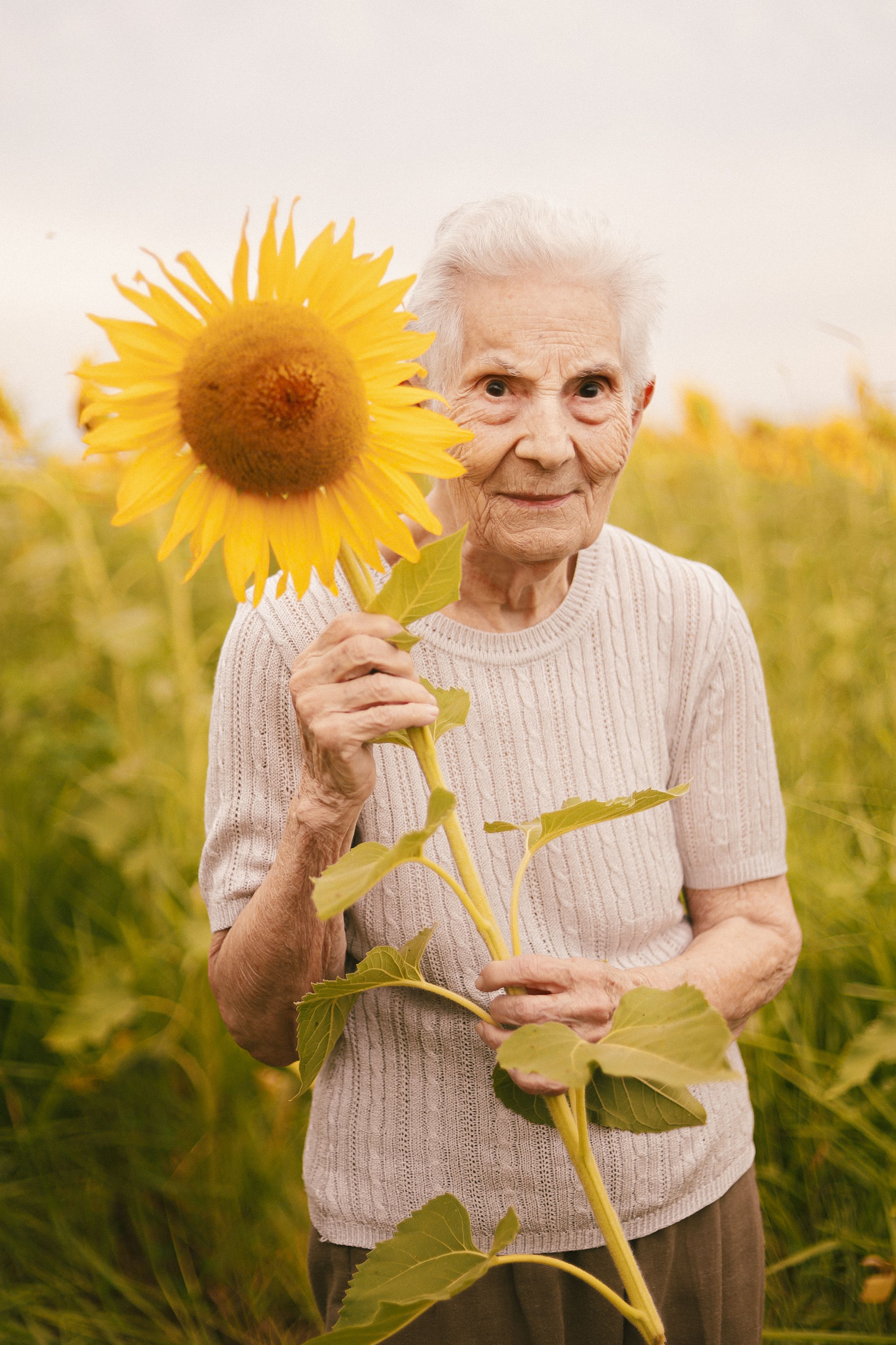 Senior woman joyfully holding a sunflower in a vibrant field during summer, expressing warmth and nostalgia.