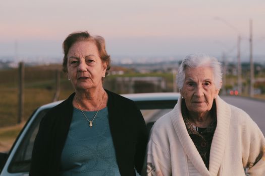 Two senior women standing together outdoors by a car during sunset.