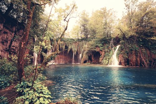 Free Serene waterfall scene in a vibrant forest with clear blue waters, offering a picturesque natural retreat. Stock Photo