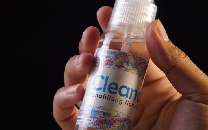 Close-up of a hand holding a clear spray bottle labeled 'Cleaner' against a dark background.