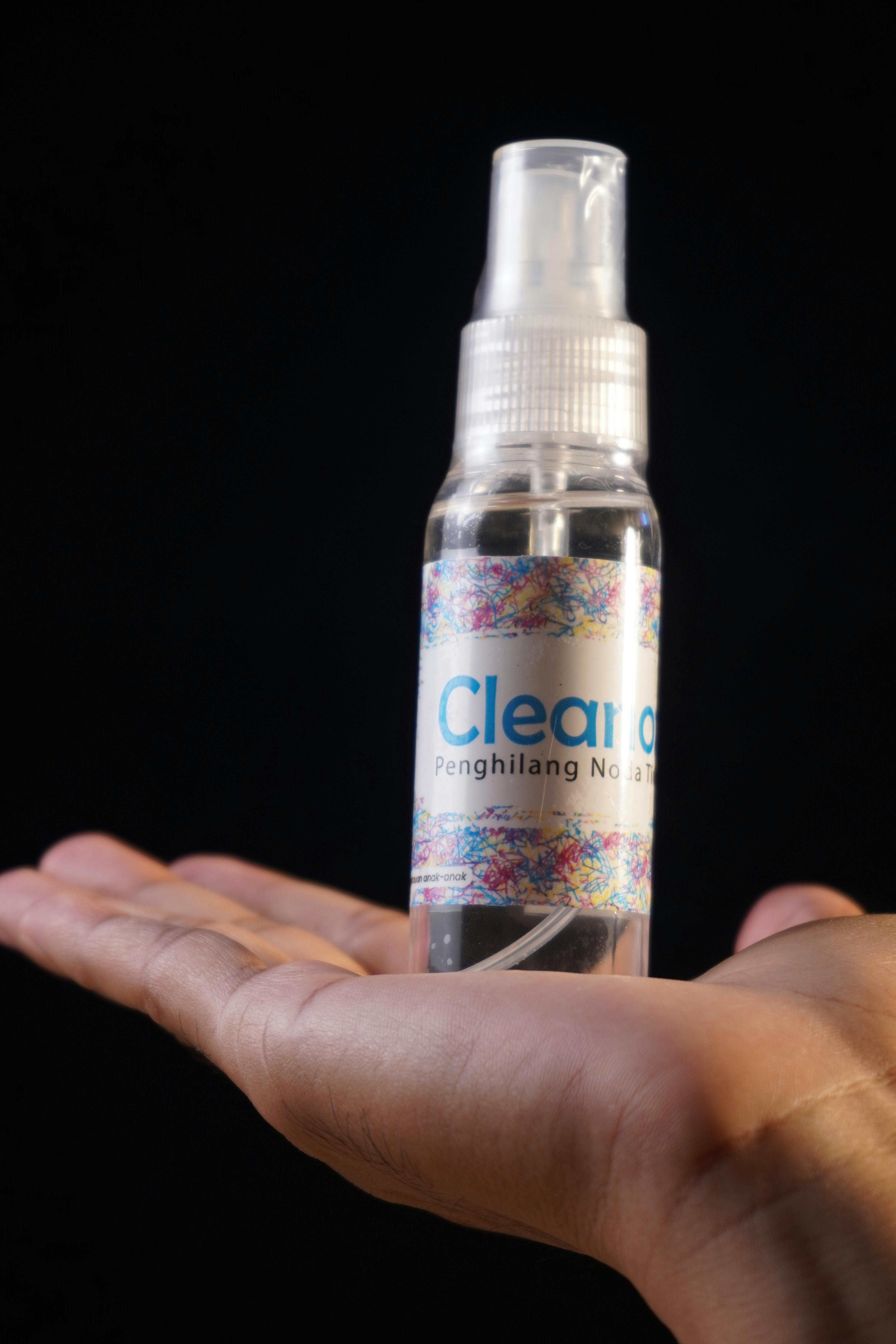A clear spray bottle labeled with text is balanced on an open hand against a dark background.