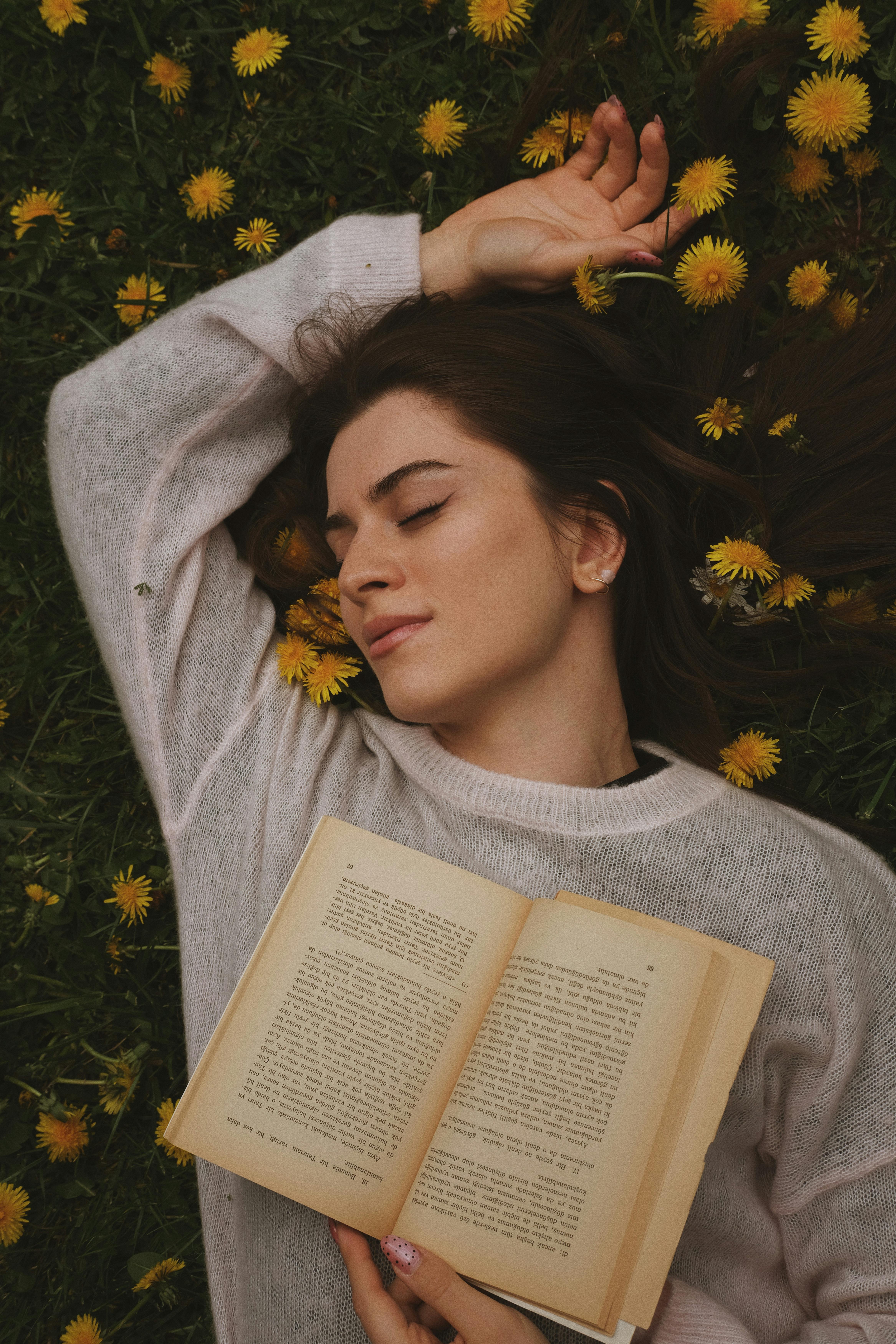 A serene woman lies in a field of dandelions, holding an open book and embracing tranquility.