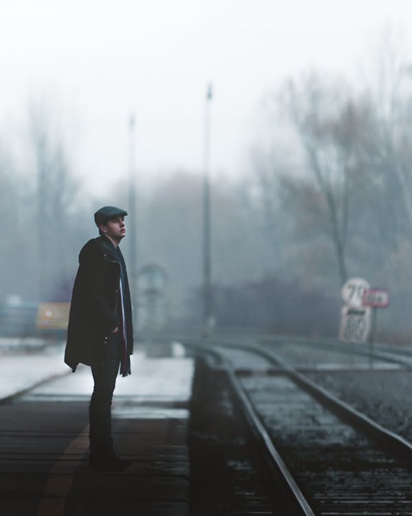 Side view full body unemotional male wearing outerwear standing with hand in pocket on empty railway station platform while looking away thoughtfully during winter day