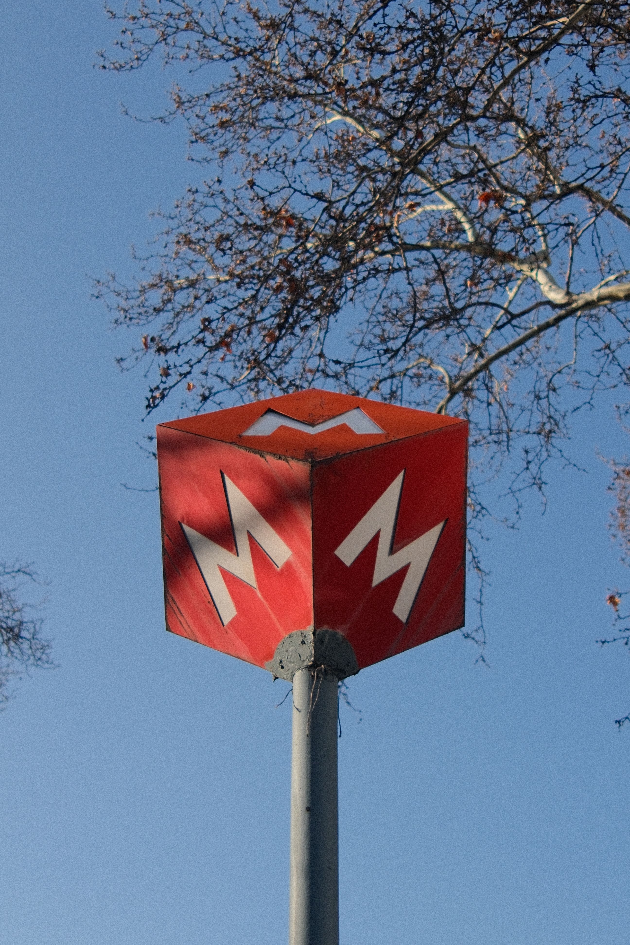 Red metro sign with bold white 'M' against a clear sky and tree branches.