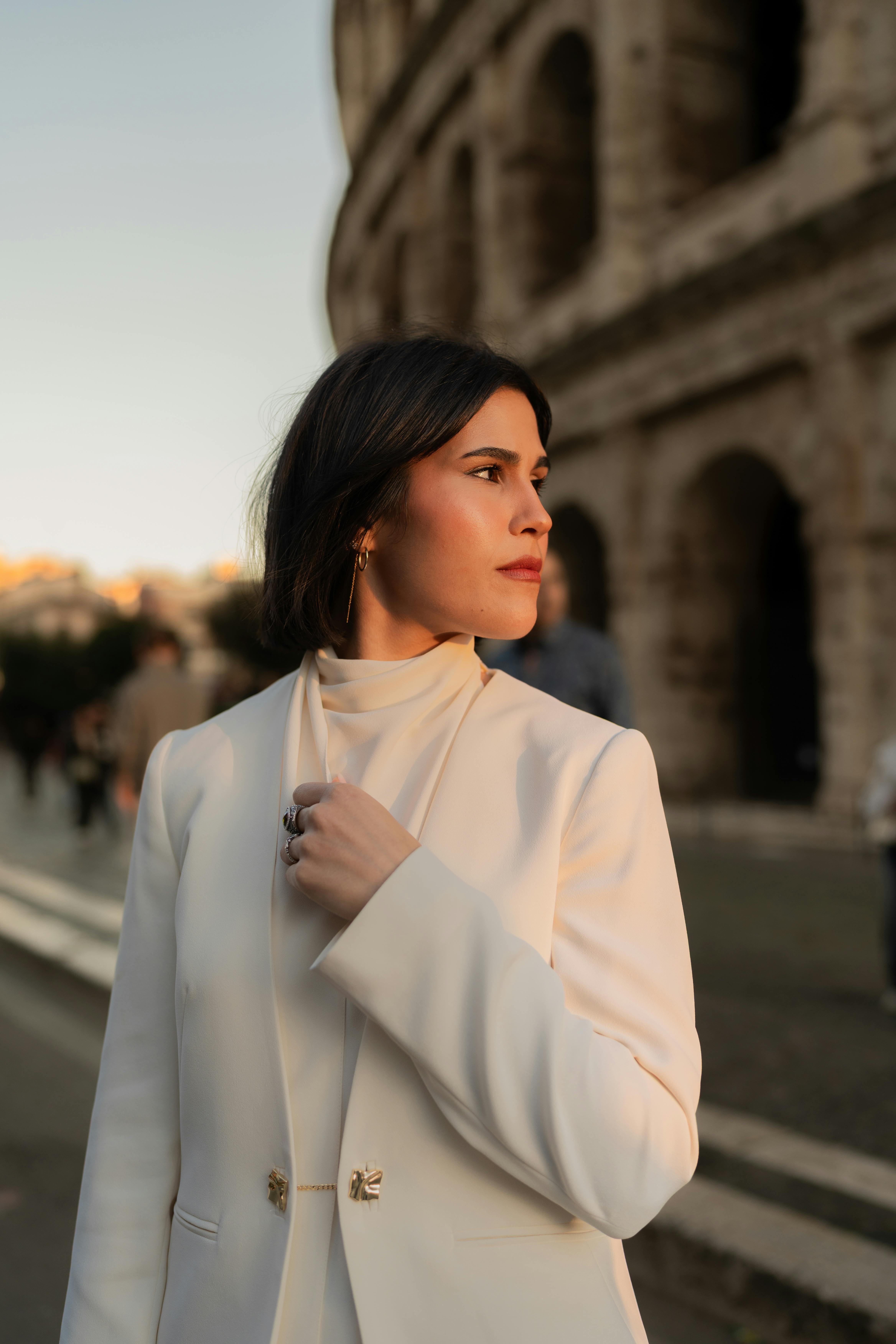 Stylish woman in white attire gazes thoughtfully near Rome's Colosseum at sunset.