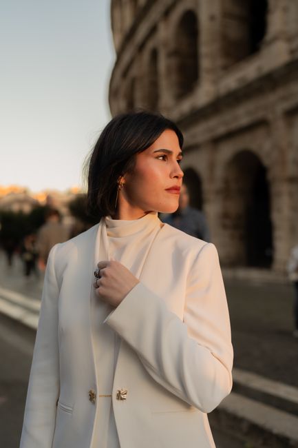 Stylish woman in white attire gazes thoughtfully near Rome's Colosseum at sunset.