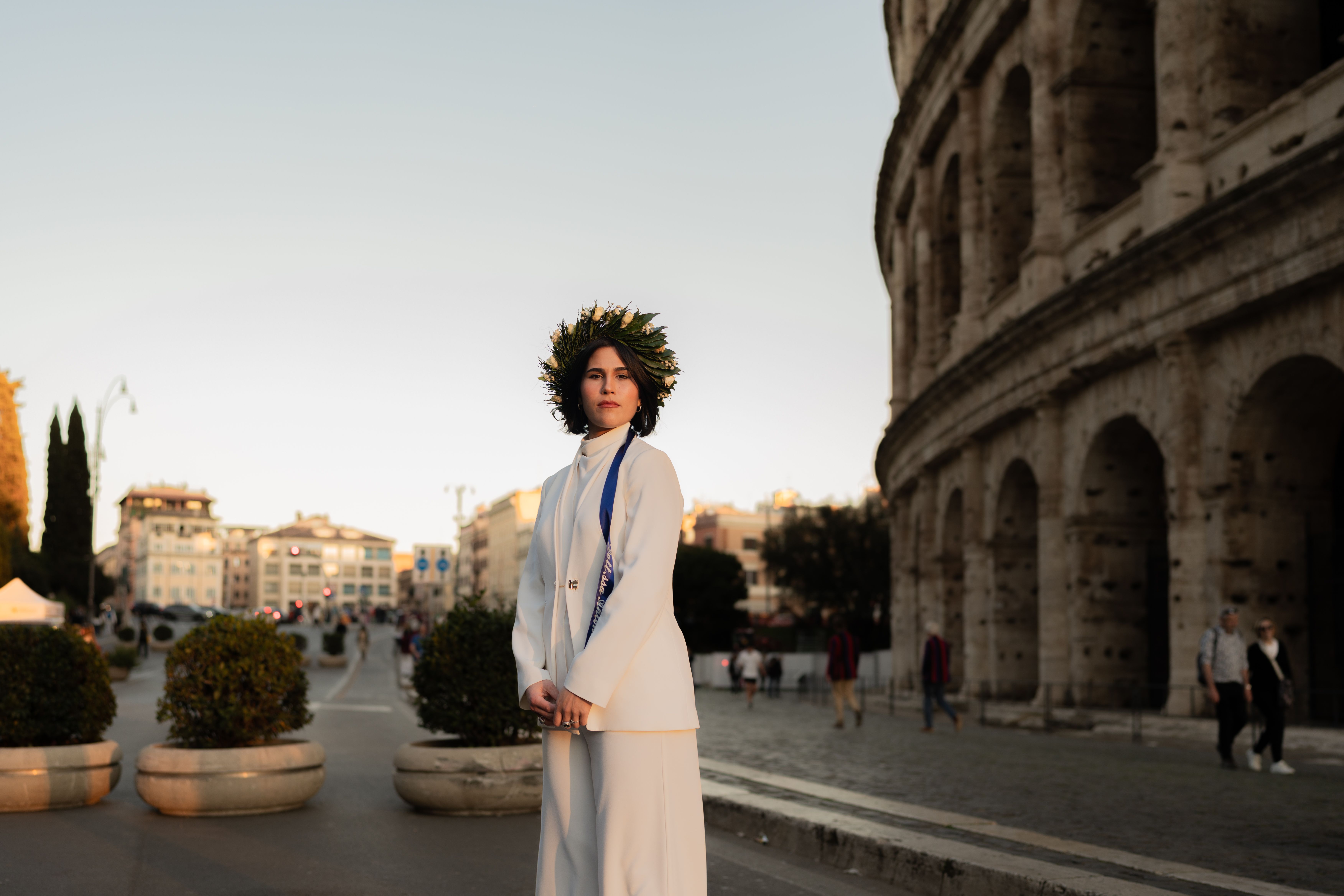 Stylish young woman poses in front of Rome's iconic Colosseum at sunset wearing a white outfit.