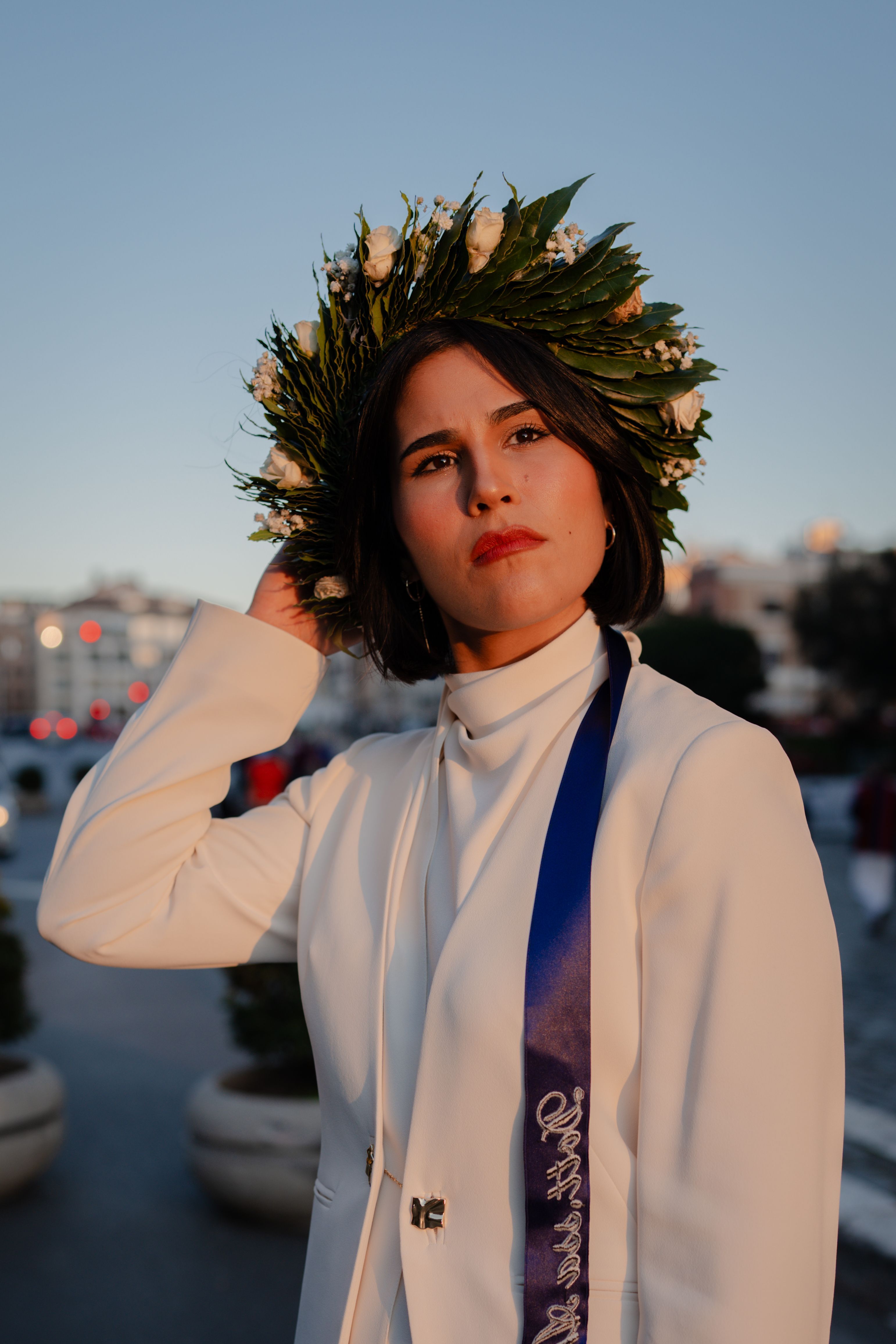 A woman in a white suit and laurel wreath poses confidently in an outdoor setting during sunset.