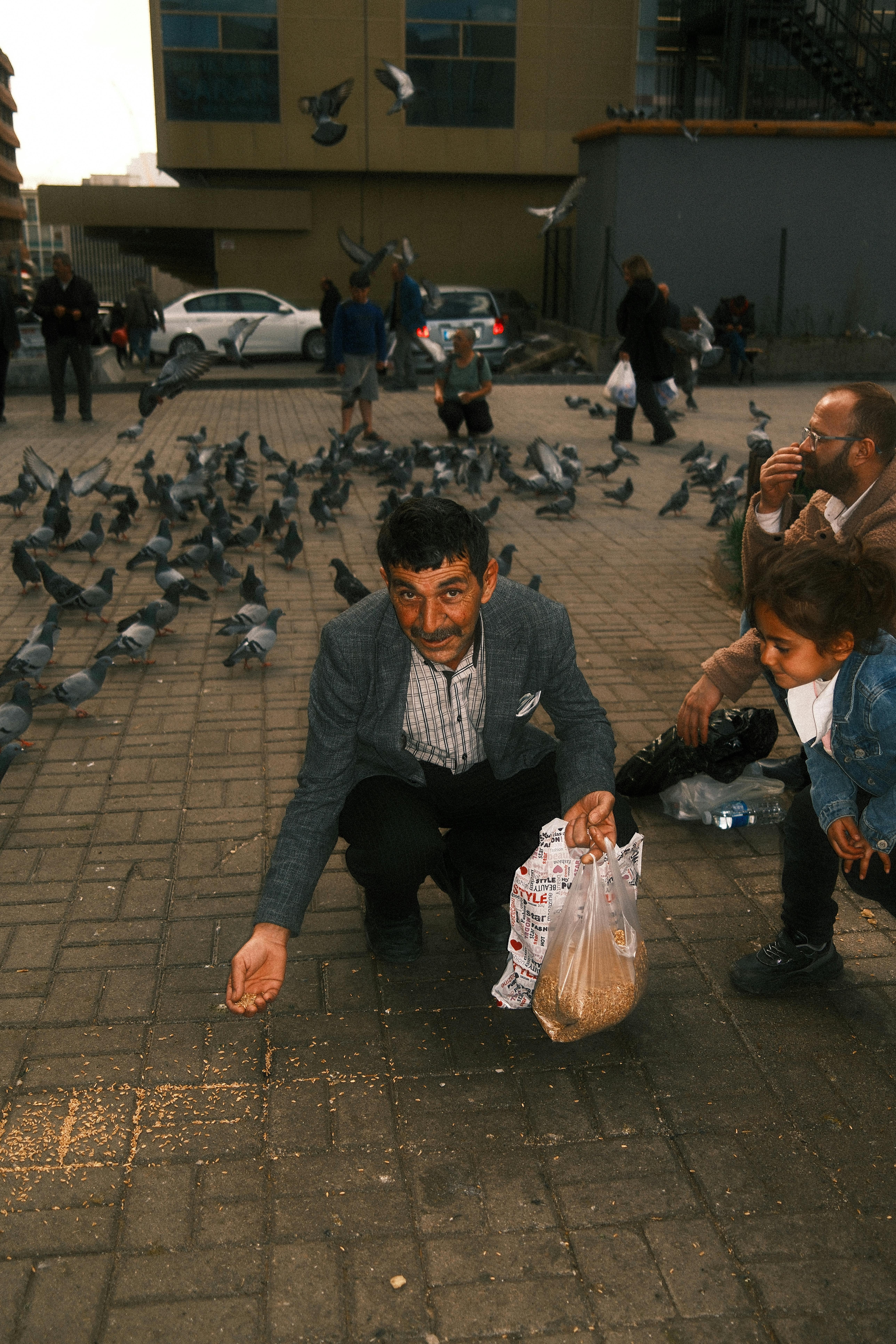 Bezpłatne Mężczyzna kuca na chodniku w mieście i karmi gołębie, tworząc ożywioną miejską scenerię. Zdjęcie z galerii