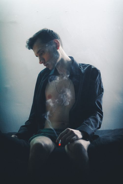 Free A young man sits indoors, smoking in a dimly lit room, reflecting a moody and contemplative atmosphere. Stock Photo