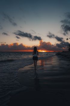 A woman walks along a serene beach during a vibrant sunset, reflecting on the water.
