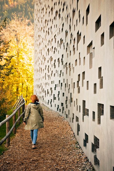 A woman in a green coat walks beside a unique, perforated wall with autumn trees in the background.