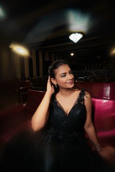 Portrait of a woman in a black dress, seated in a lavish indoor setting with chandeliers.
