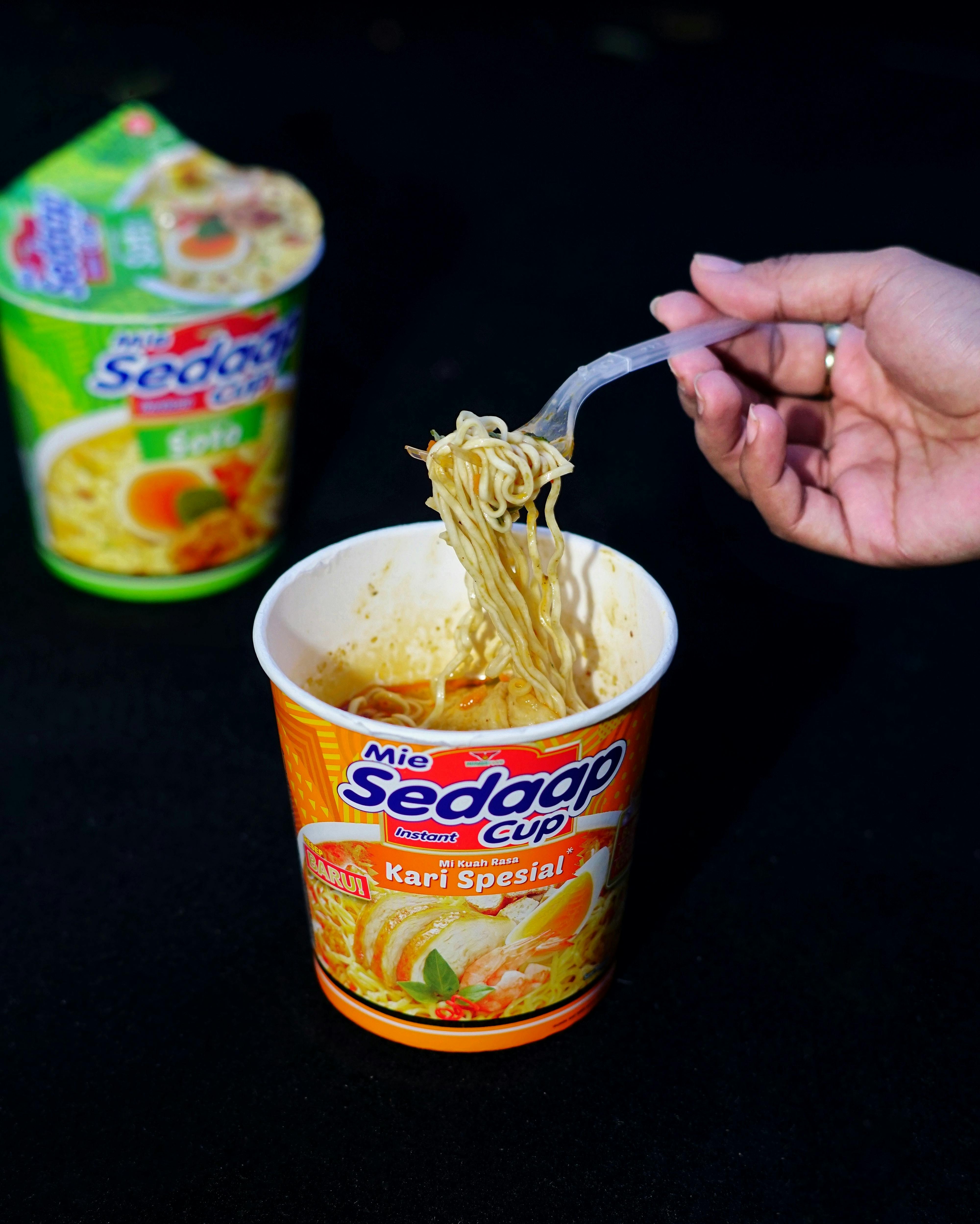 A hand lifts noodles from a flavorful curry spiced cup, ready to eat.