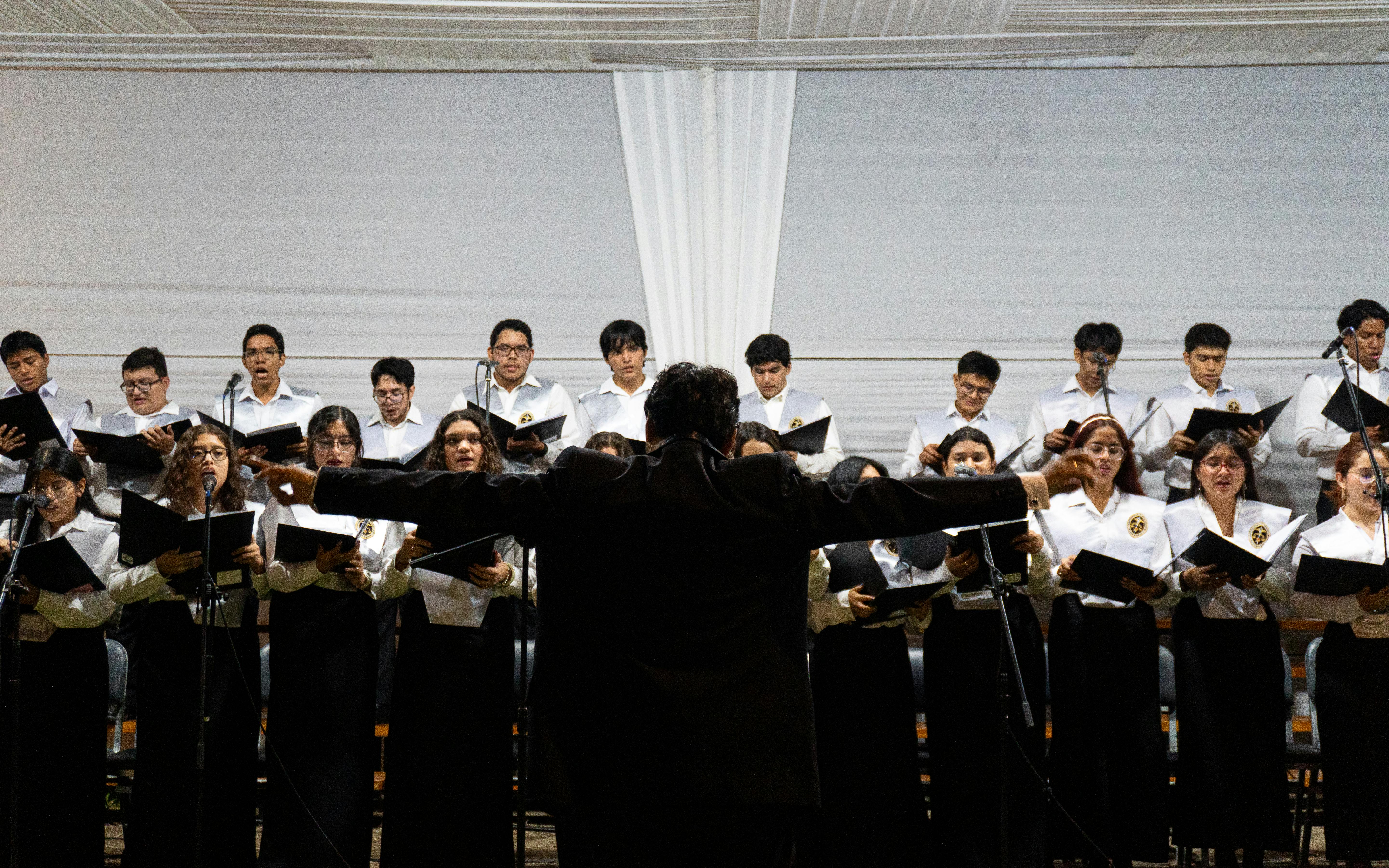 University choir performing at an indoor ceremony with conductor leading the ensemble in Piura, Peru.
