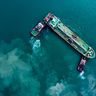 An aerial, top-down view of an oil tanker being guided by tugboats through vibrant teal waters.