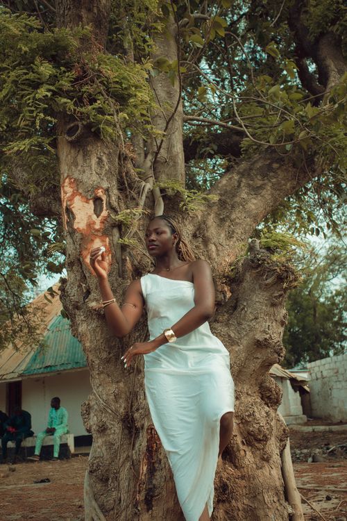 Free Graceful woman in white dress leaning against a large tree, highlighting natural beauty in Nigeria. Stock Photo