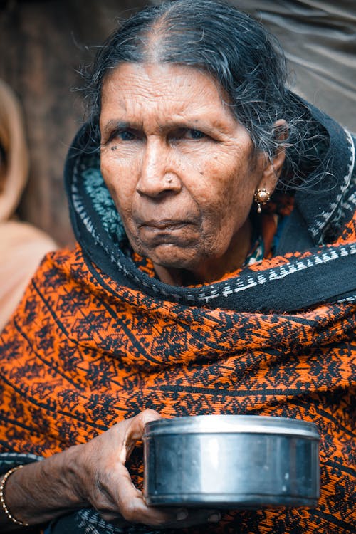 Senior Asian female in authentic national clothes with steel bowl standing on street market and looking at camera