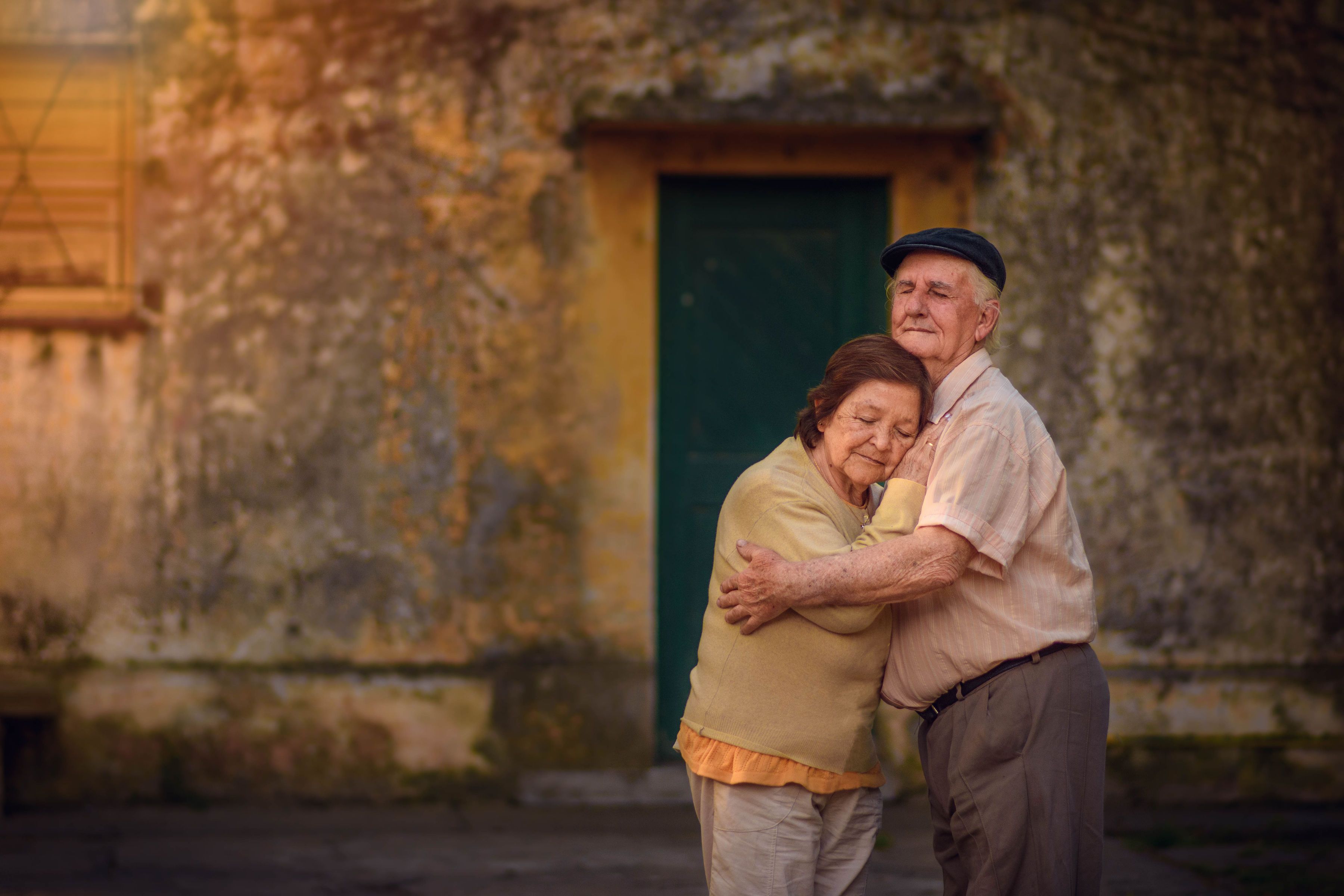 Charming elderly couple embracing warmly in front of a rustic building wall.