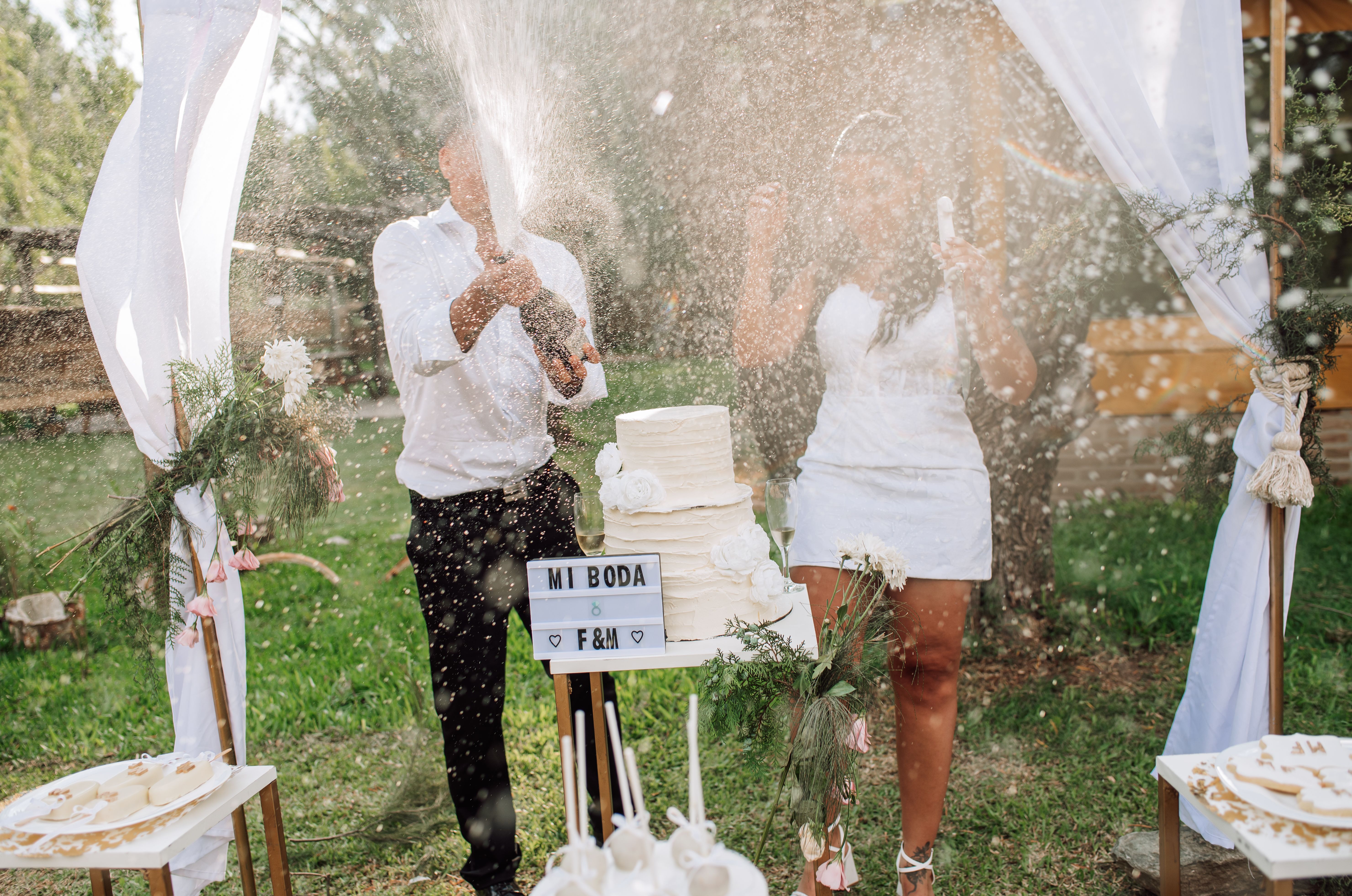 A couple celebrates their wedding outdoors with cake cutting and champagne popping under a floral arch.