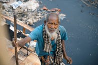 From above cheerless elderly Asian male in worn out clothes standing near dirty polluted river full of rubbish in poor district and looking at camera