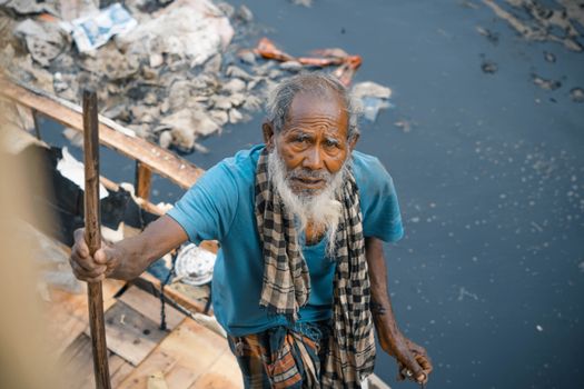 From above cheerless elderly Asian male in worn out clothes standing near dirty polluted river full of rubbish in poor district and looking at camera