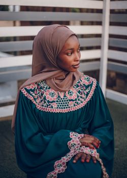 Elegant portrait of a woman wearing a hijab and traditional embroidered dress, sitting outdoors.