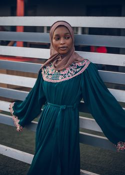 A fashionable adult woman in a detailed dress posing gracefully outdoors near a modern fence.