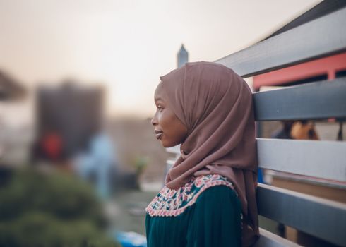 A young woman in a hijab thoughtfully leans against a bench outdoors, in a serene and soft focus background.